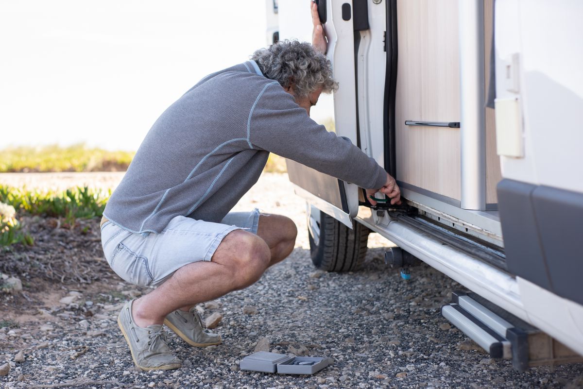 Man crouching beside an open camper van door working on the entry step with a hand tool