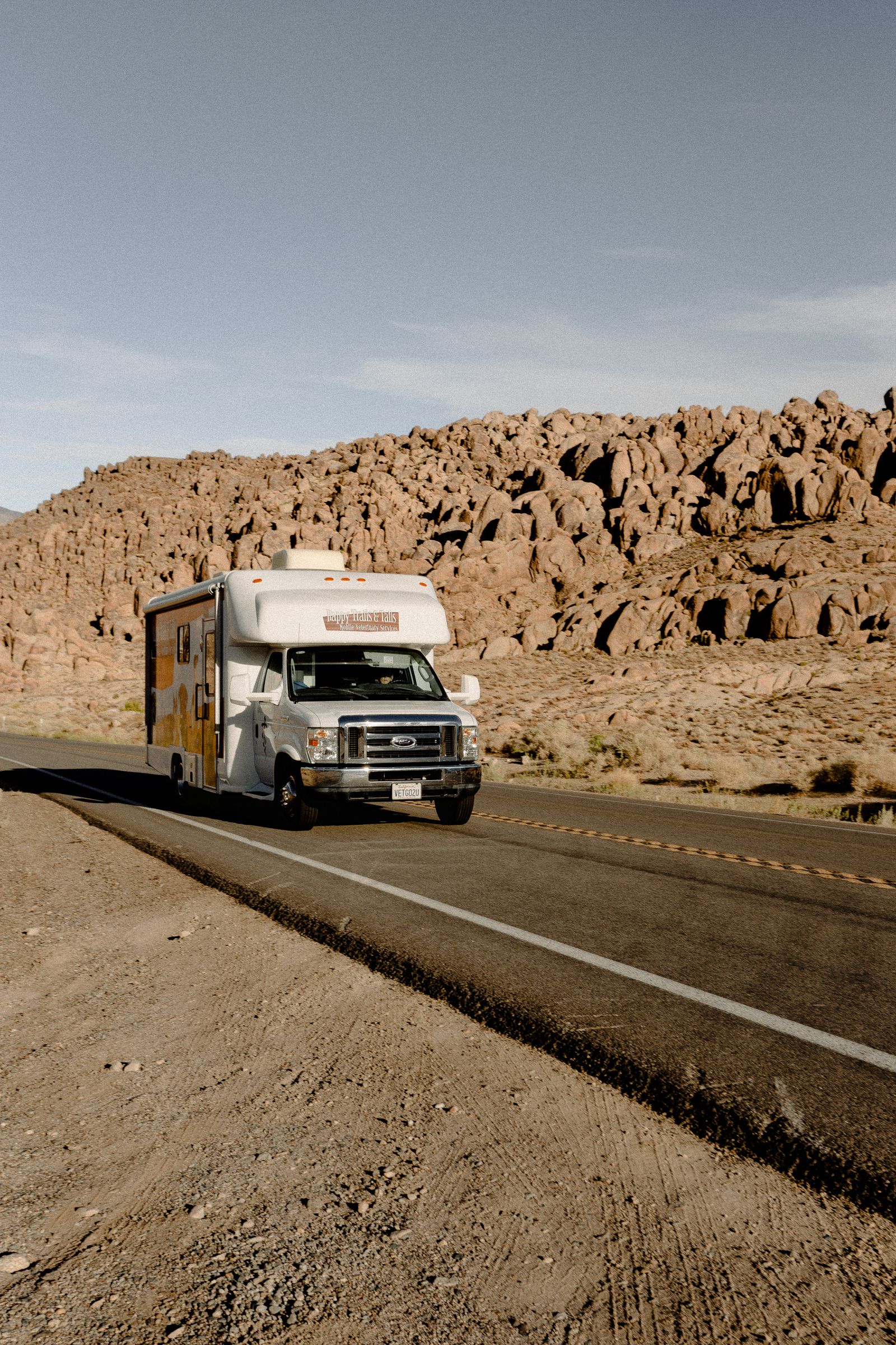 Class C motorhome driving a two-lane desert highway past rocky hills under bright sun