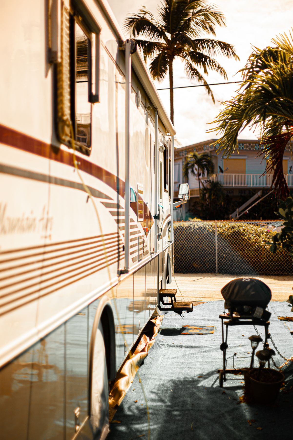 Side of a large Class A motorhome parked on an RV resort site with palm trees and a patio mat in warm afternoon light